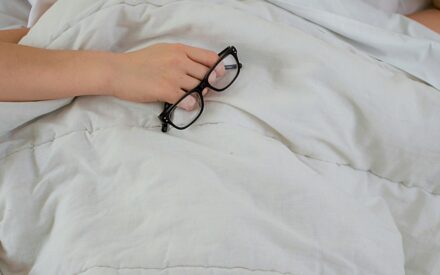 Woman holding eyeglasses in bed trying to sleep with her head under a pillow