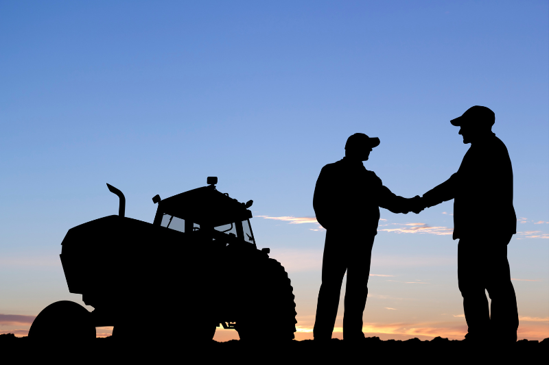 Two farmers shaking hands in a field next to a tractor
